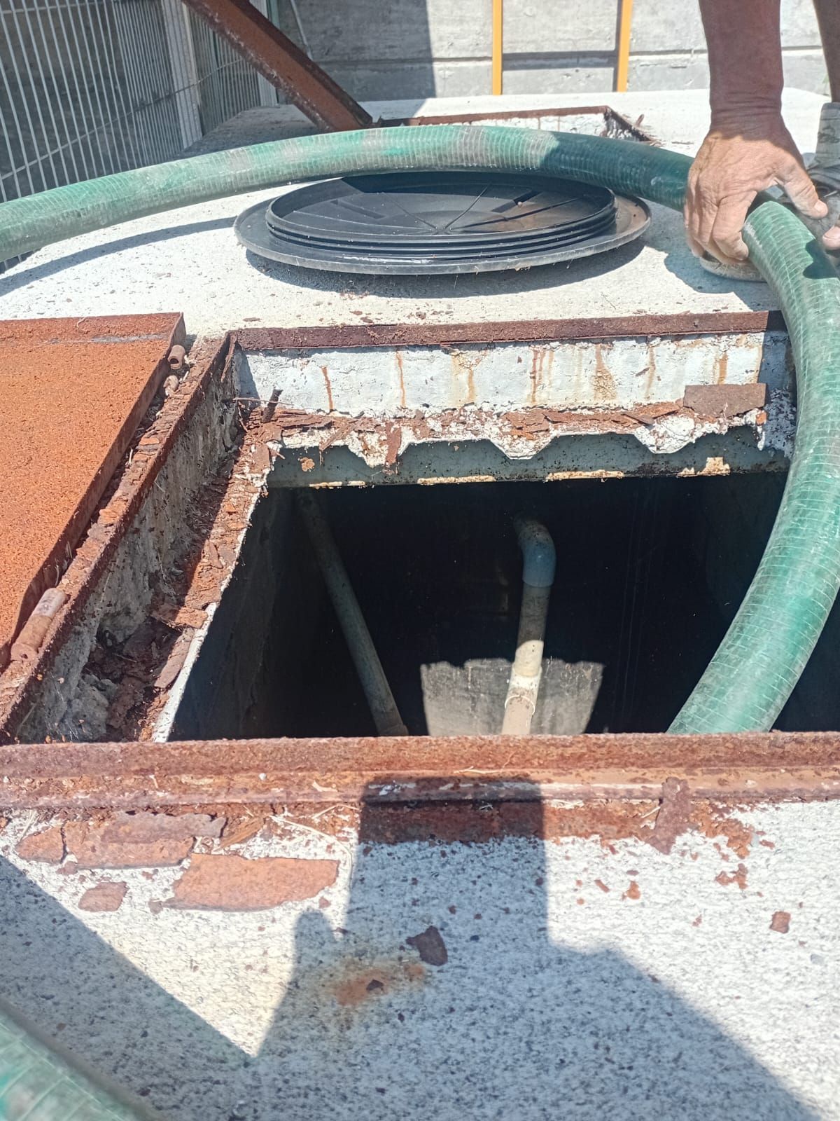 Hombre sosteniendo una manguera, mirando dentro de la abertura de un tanque oxidado, con una pala adentro.