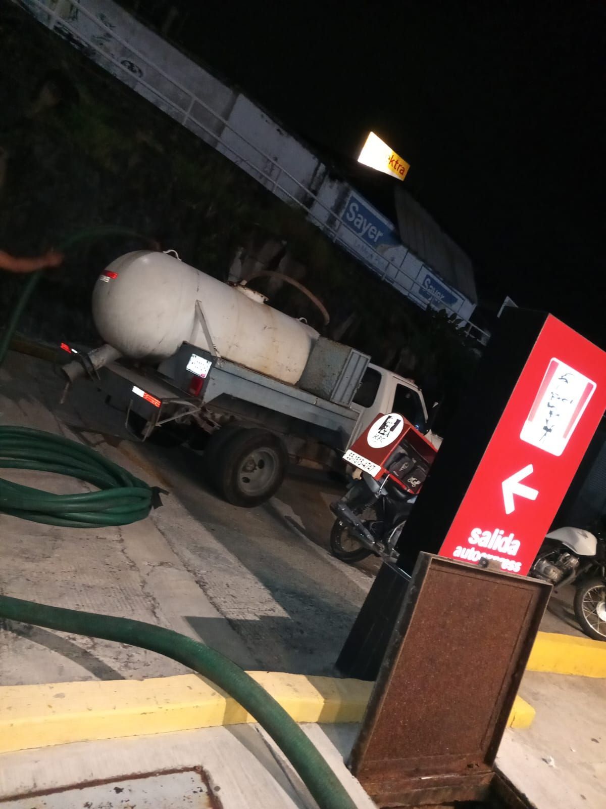 Camión con gran tanque de propano en una gasolinera, manguera en el suelo, cartel con una flecha.