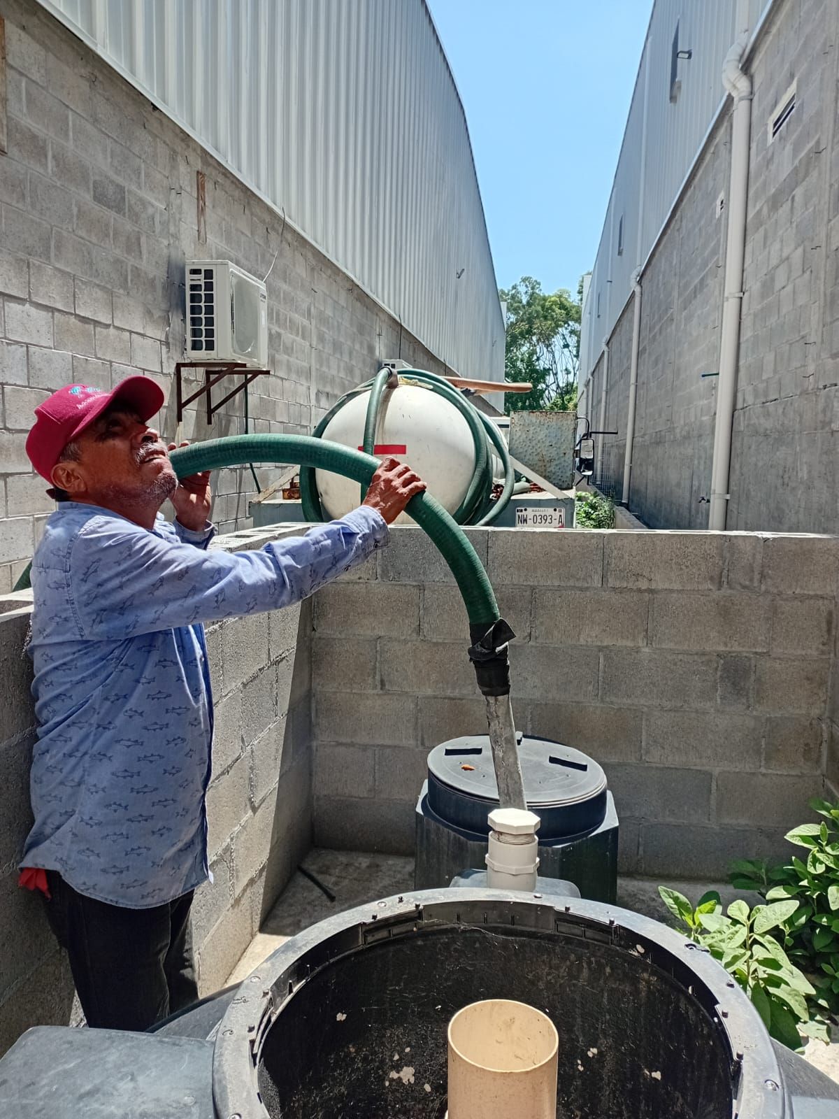 Un hombre sostiene una manguera y mira cómo el agua fluye hacia un gran tanque afuera, entre edificios, bajo un cielo azul.
