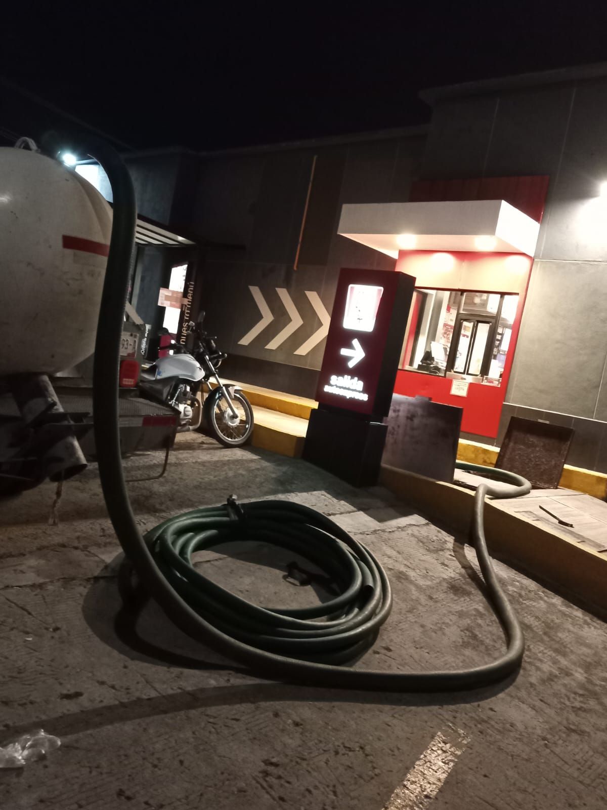 Una manguera de camión cisterna enrollada sobre el pavimento cerca de un drive-thru y una motocicleta estacionada por la noche.