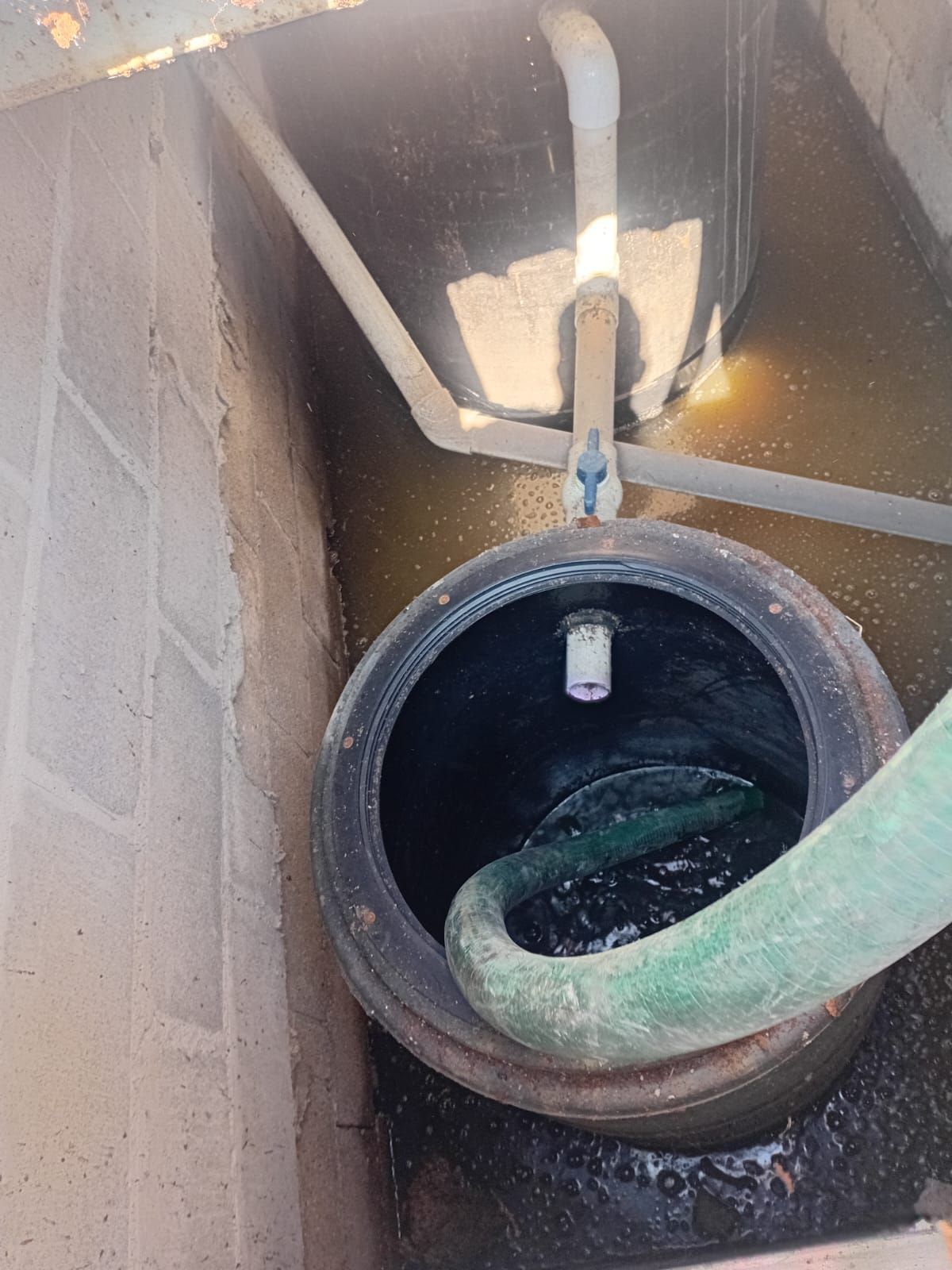 Pozo de hormigón abierto que contiene un recipiente negro con una manguera y agua, y un tanque negro más pequeño, con tuberías de conexión.