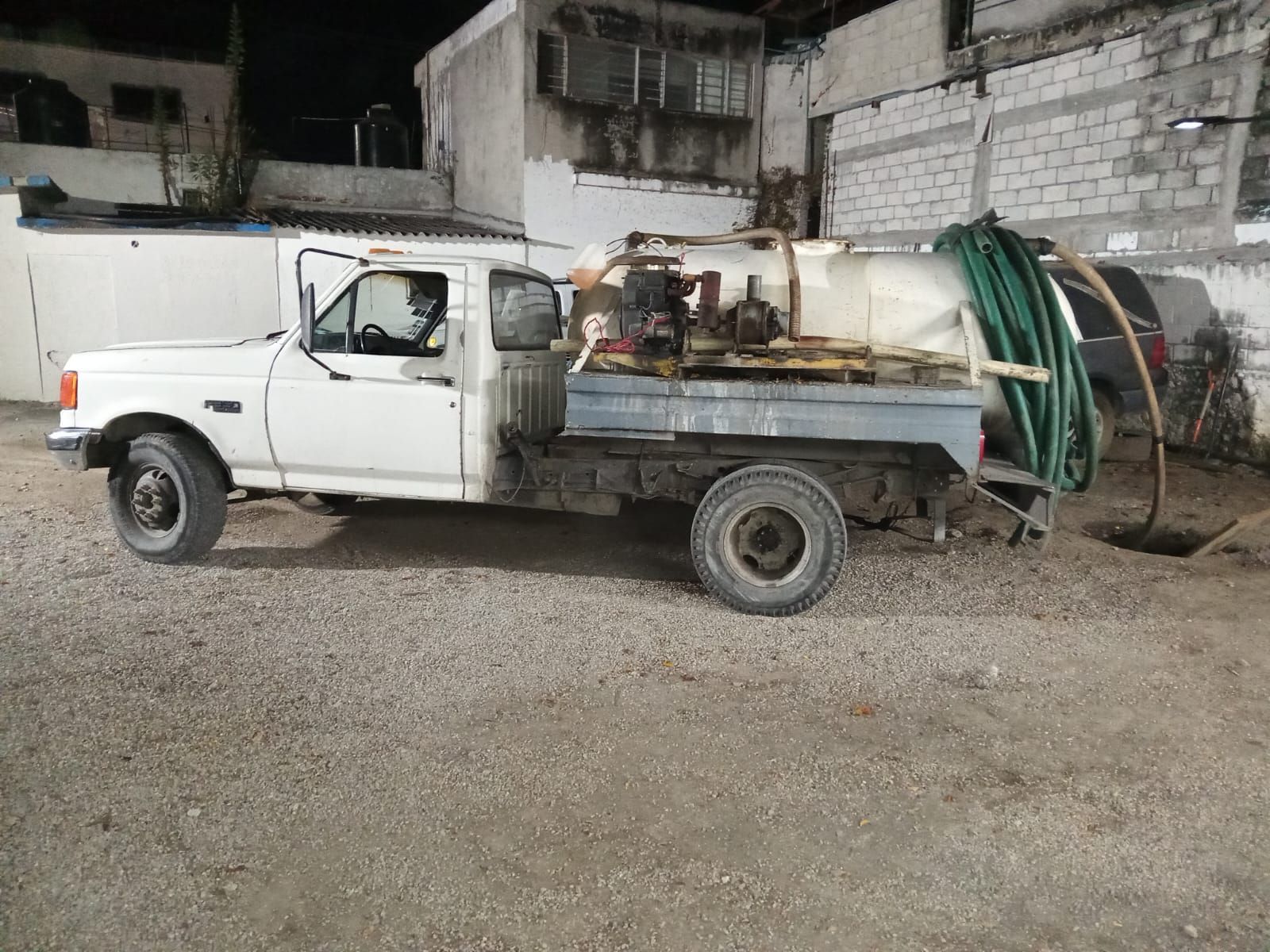 Camión blanco con cisterna en la plataforma, estacionado sobre grava. Una manguera verde está conectada a la cisterna.
