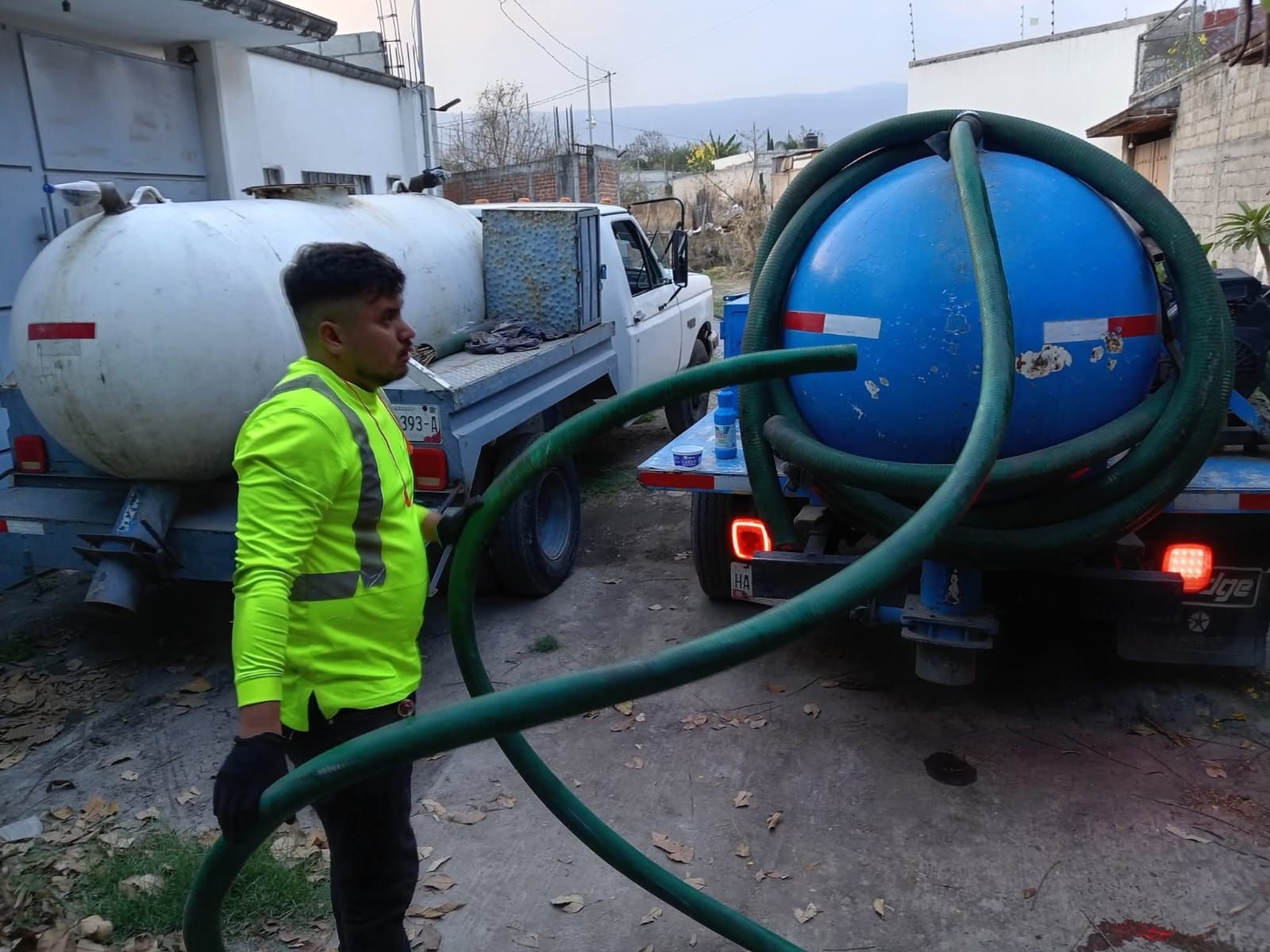Hombre con mangueras cerca de dos camiones cisterna, uno azul y otro blanco, al aire libre.