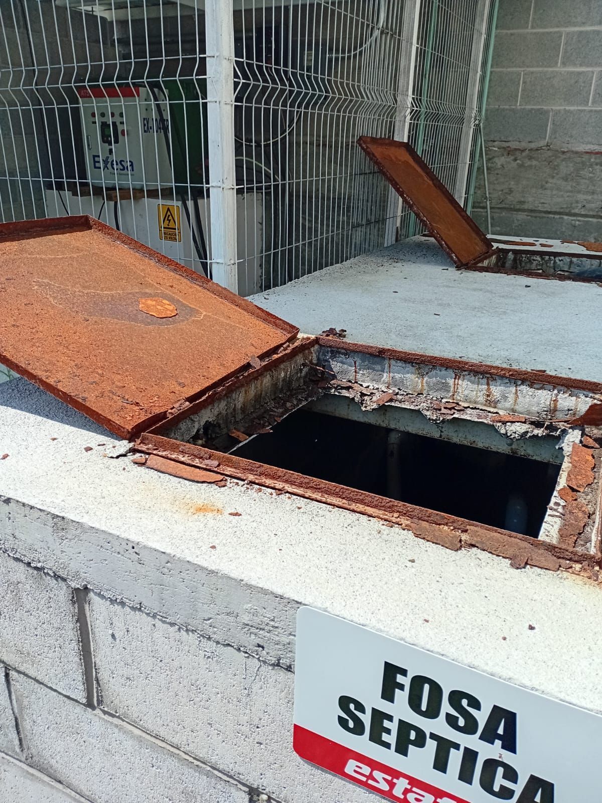 Tanque séptico abierto con tapas oxidadas. Letrero de advertencia: