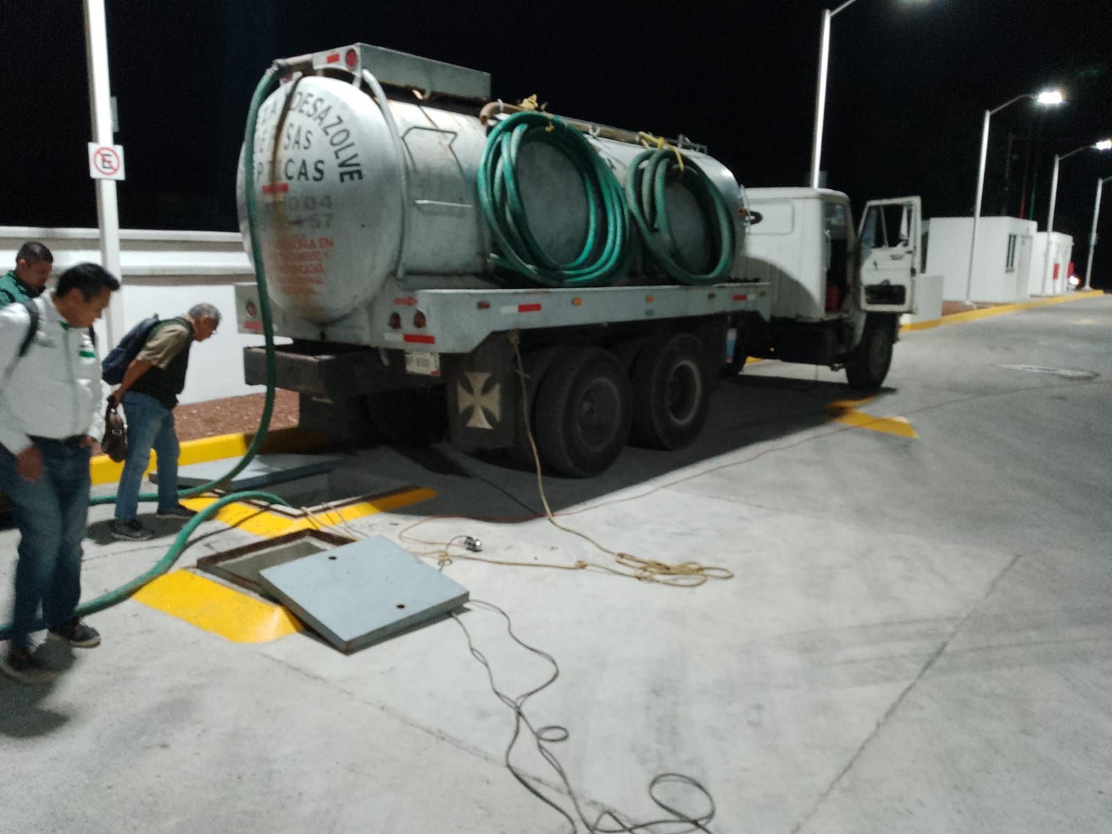 Un camión cisterna está estacionado cerca de un desagüe de hormigón. Hay hombres cerca y las mangueras están conectadas.