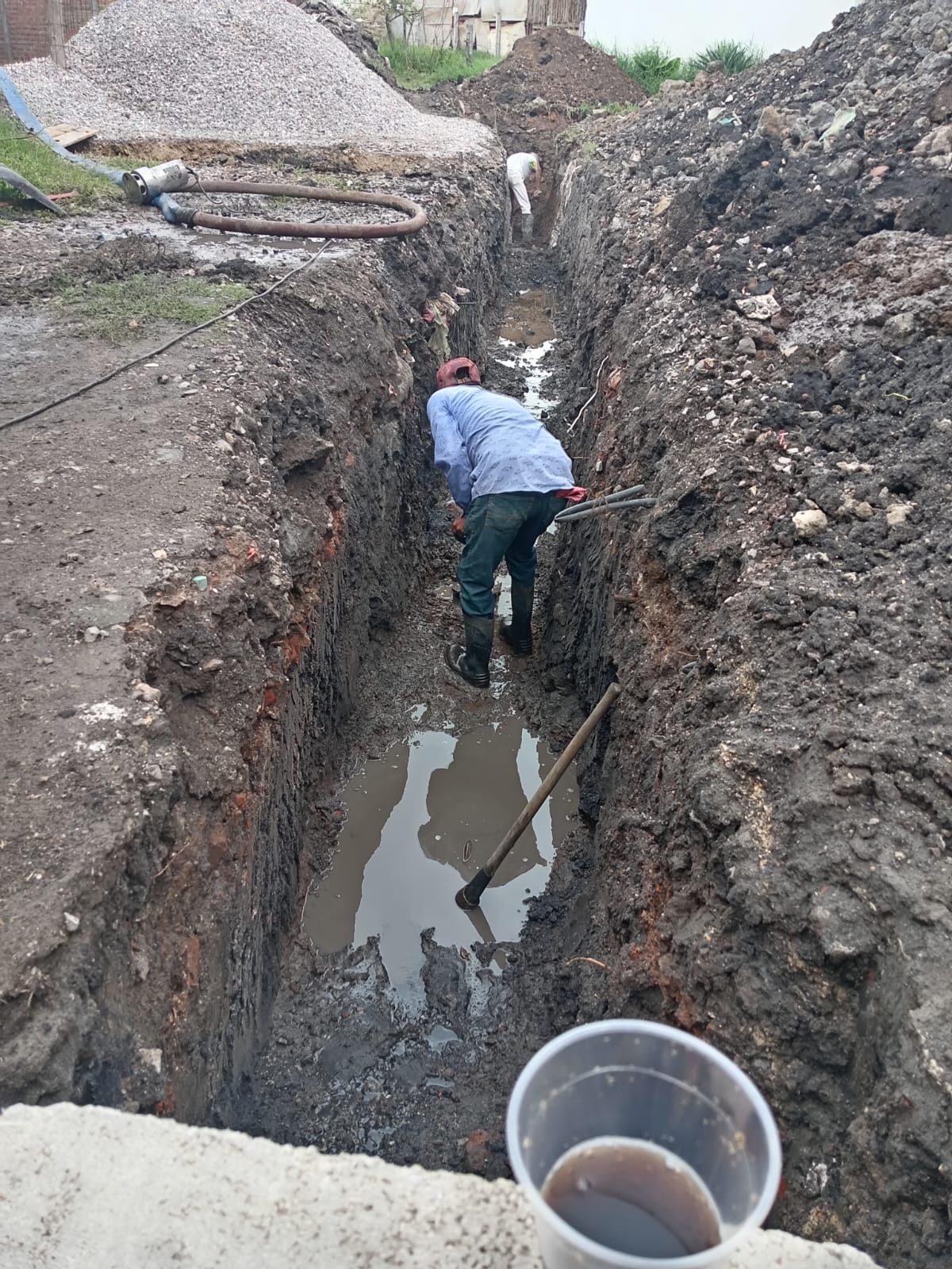 Persona paleando en una zanja lodosa; pila de grava y edificio al fondo. Vaso de plástico en primer plano.