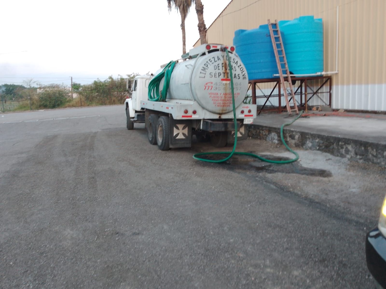 Camión cisterna llenando tanques de agua cerca de un edificio. Manguera verde, tanques azules y turquesas.