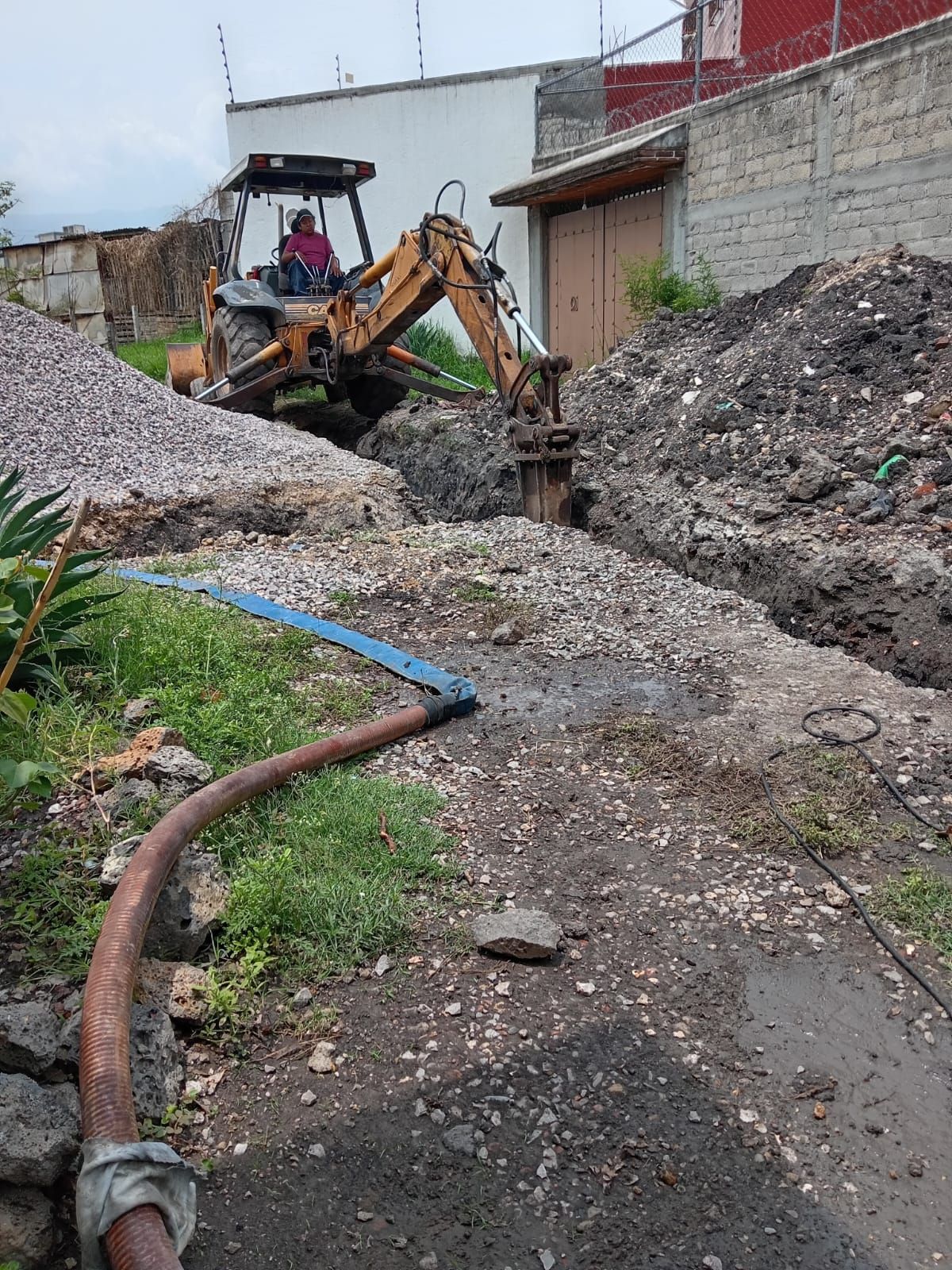 Retroexcavadora cavando una zanja cerca de un edificio. Manguera en el suelo; montones de grava y tierra.