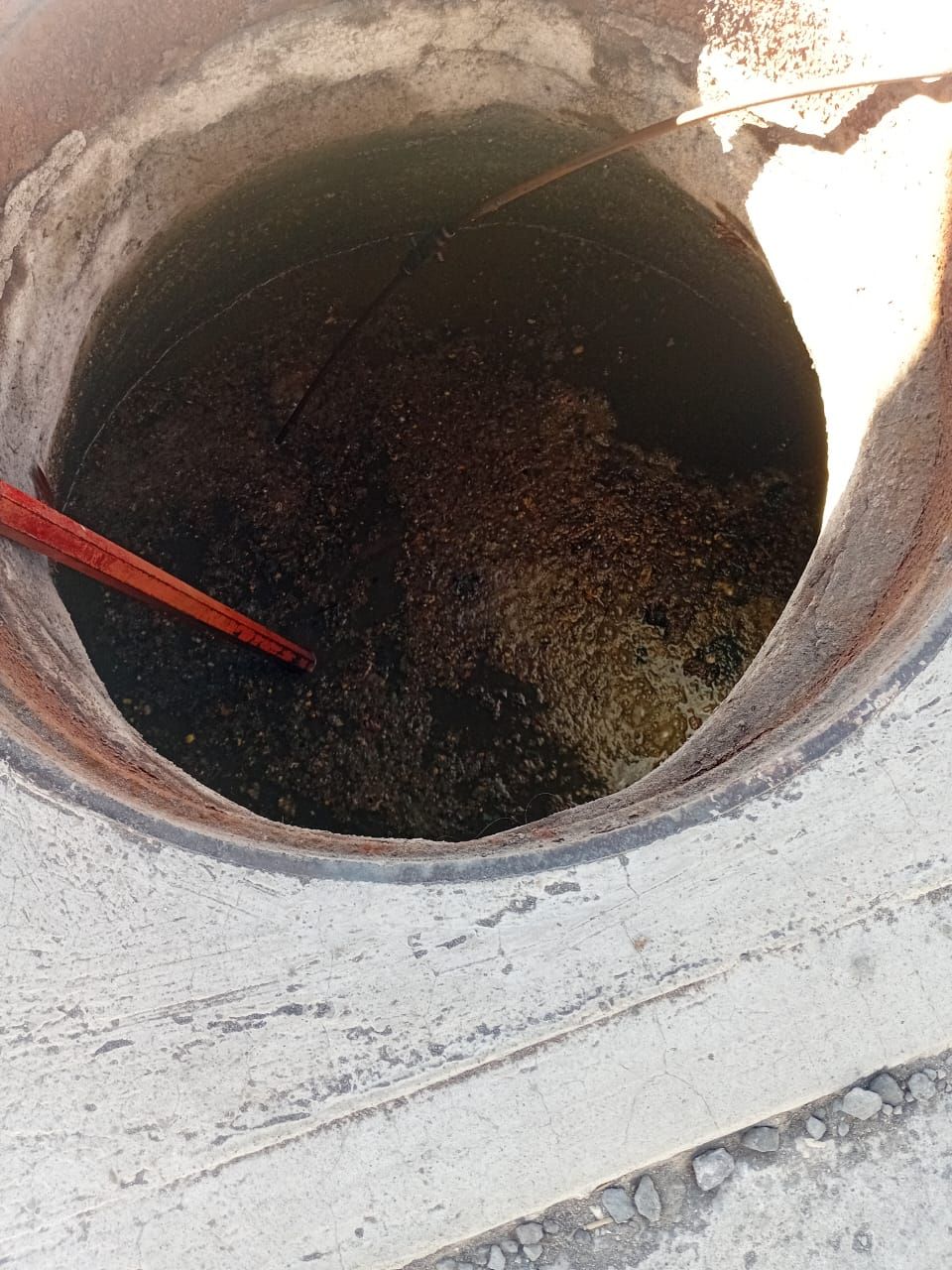 Pozo abierto lleno de escombros oscuros y líquido; hay una herramienta roja dentro.