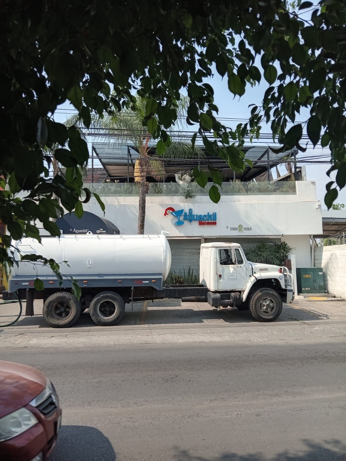 Camión de agua blanca estacionado frente a un edificio blanco con techo y un cartel de restaurante.