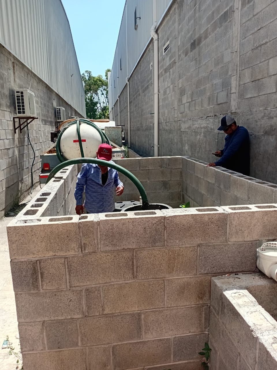 Dos hombres cerca de estructuras de bloques de hormigón, uno de ellos atendiendo un tanque con una manguera. Edificios flanquean el espacio.