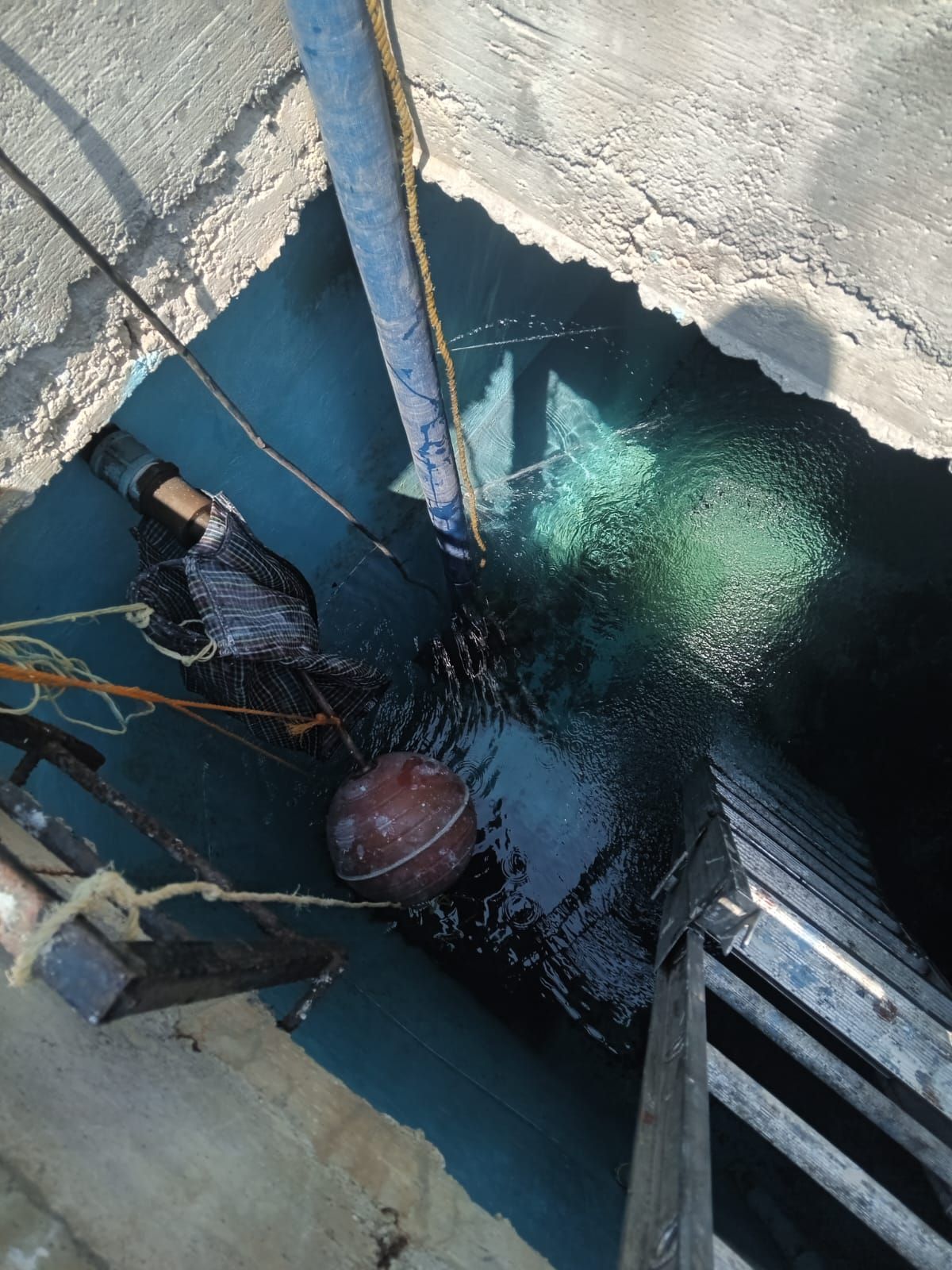 Mirando hacia abajo a un tanque oscuro lleno de agua. Se ven una manguera azul, una boya y una escalera.