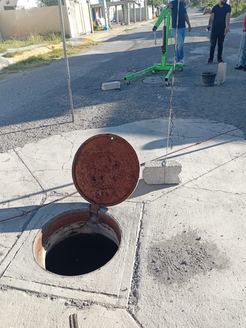 Alcantarilla abierta en una acera; tapa sujeta con soportes. Personas al fondo.
