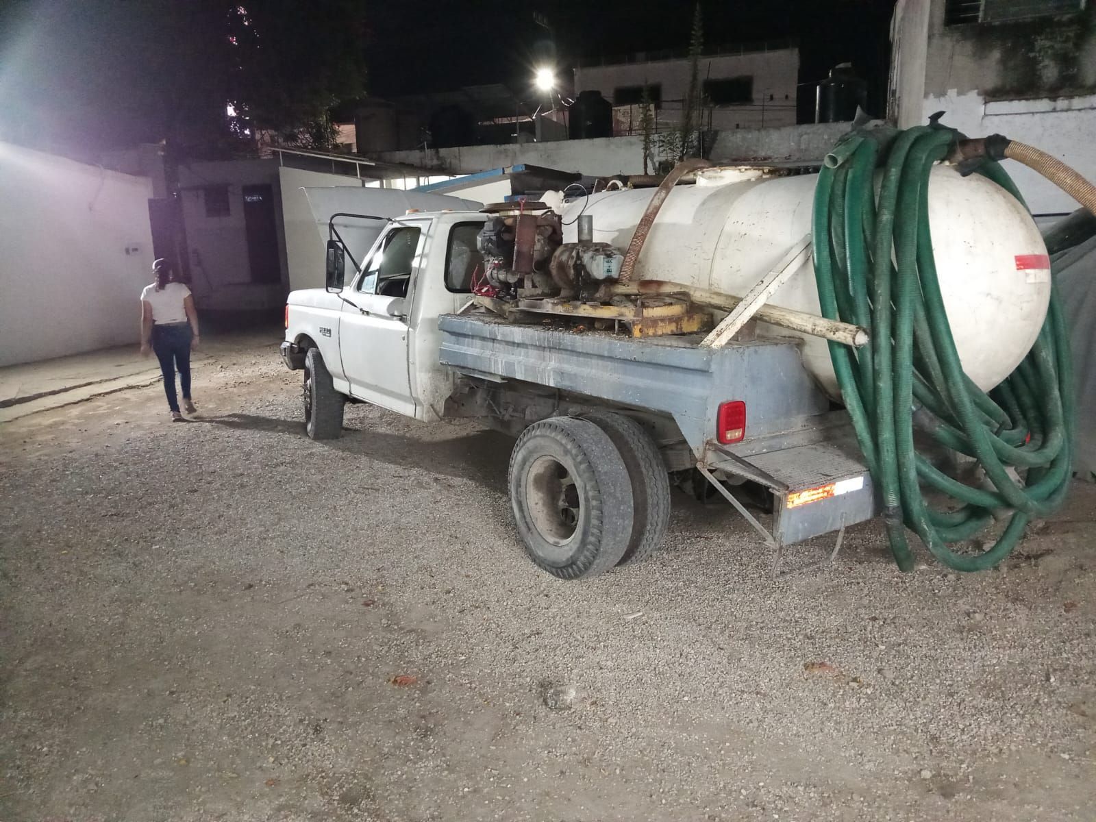 Camión séptico blanco con manguera enrollada en la parte trasera, estacionado sobre grava, una persona camina cerca.