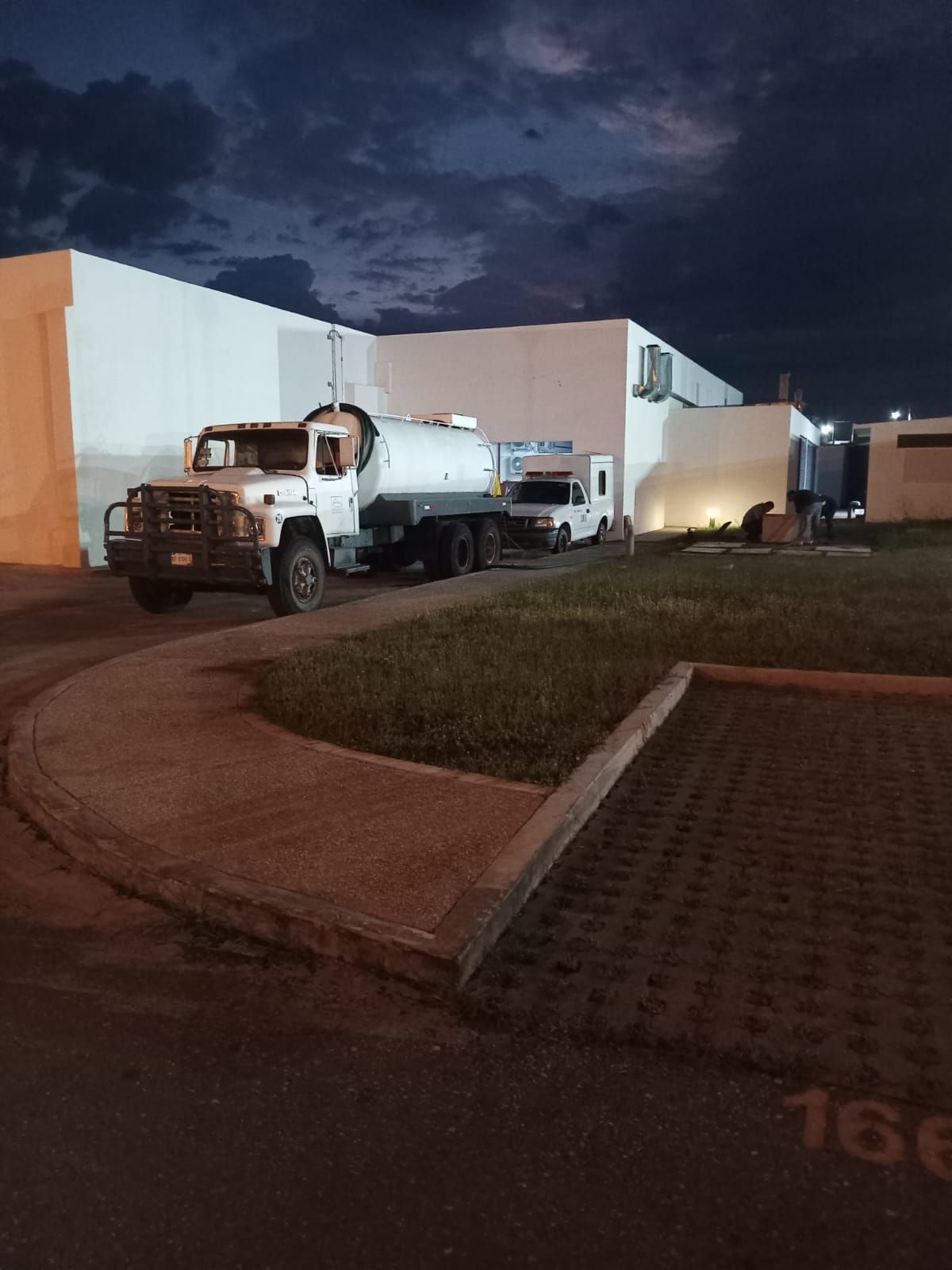 Camión cisterna blanco estacionado cerca de un edificio al anochecer, en una zona pavimentada con césped.