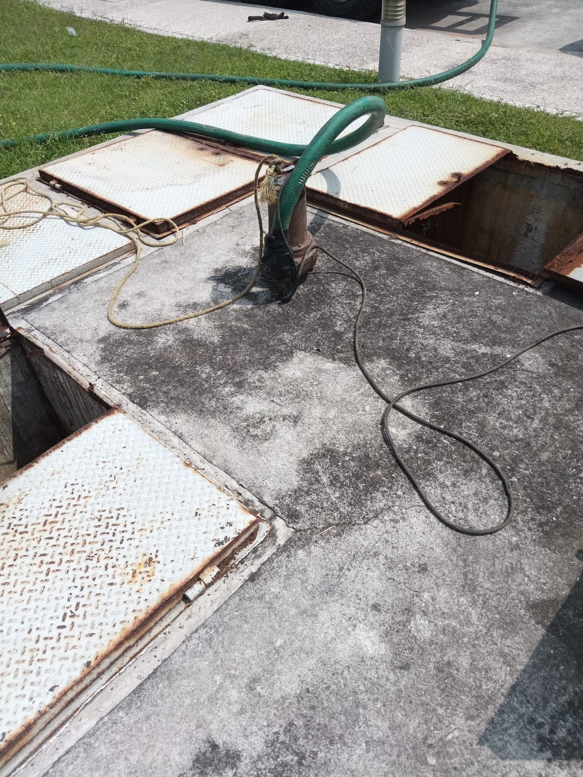 Un recinto de hormigón con tapas abiertas, una bomba en su interior y una manguera verde conectada a ella.