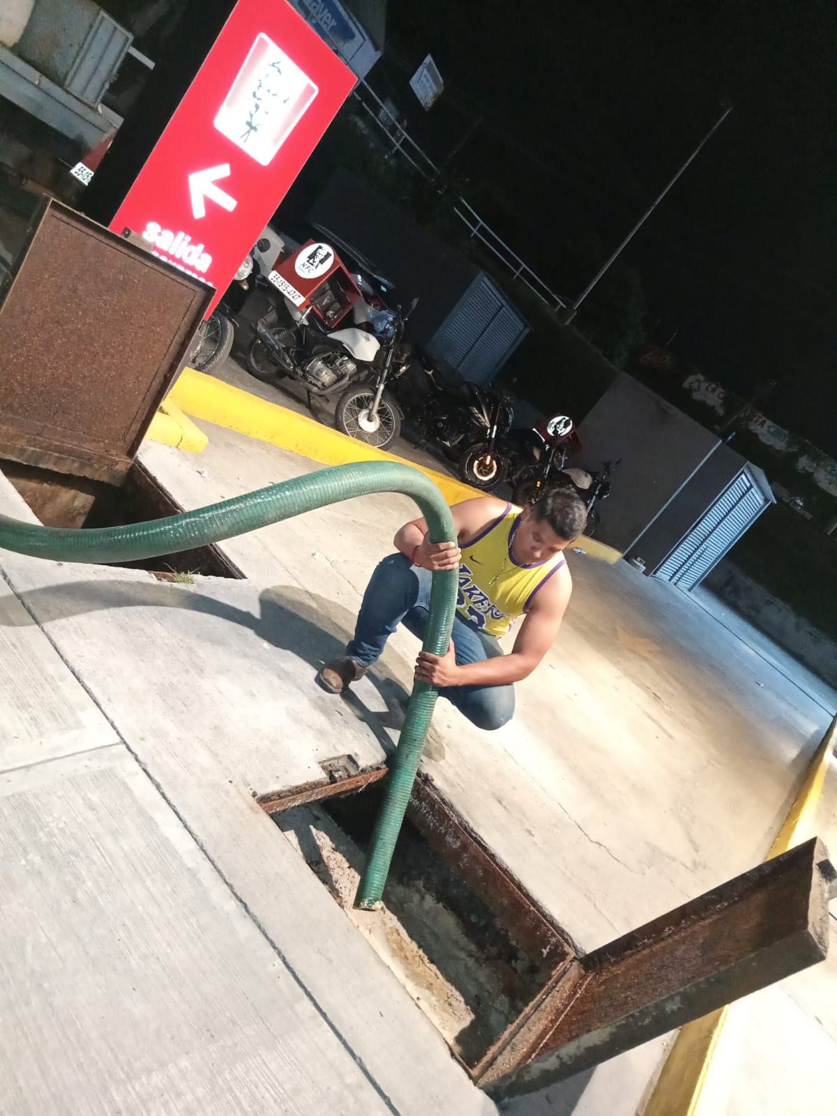 Hombre arrodillado, sosteniendo una manguera, sobre un desagüe abierto por la noche; motocicletas visibles en el fondo.