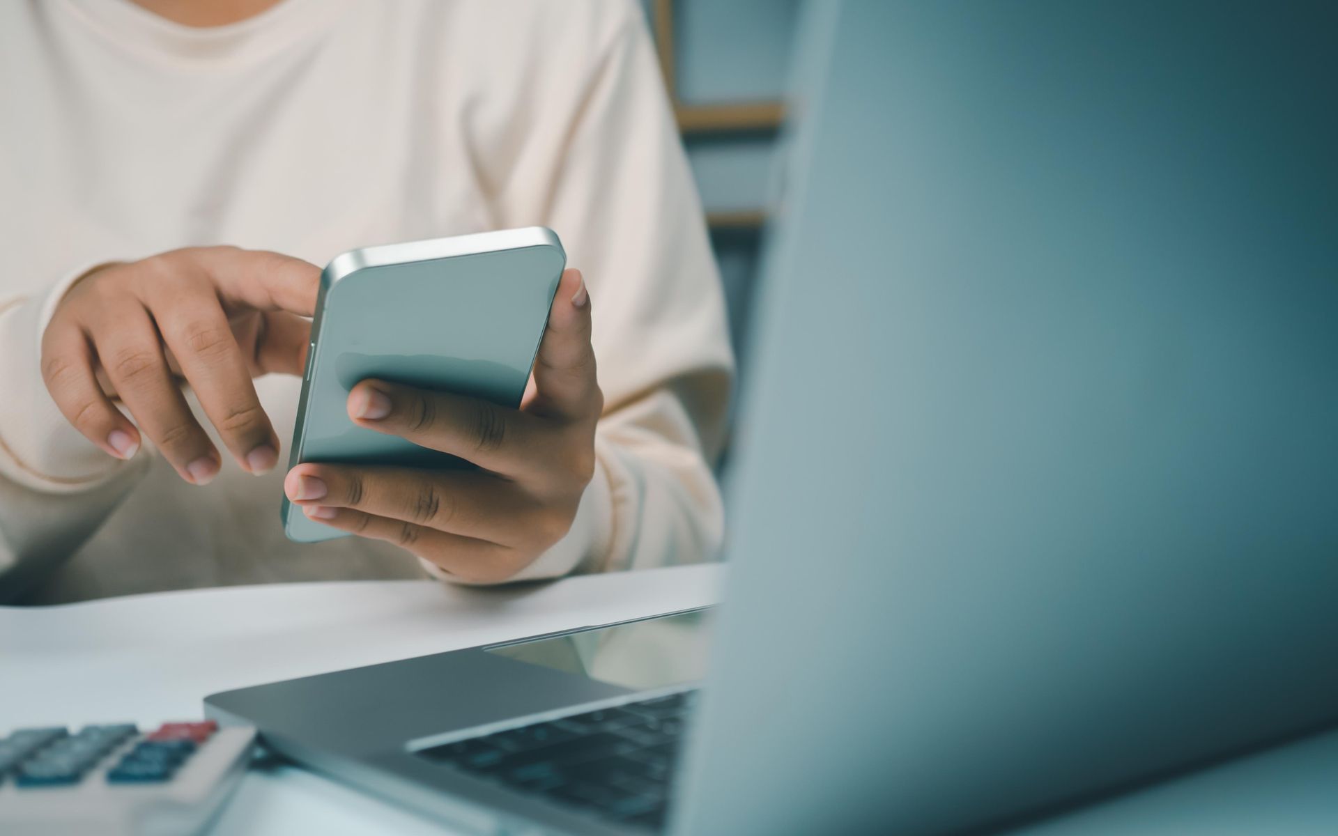 A woman is sitting at a table using a cell phone and a laptop.