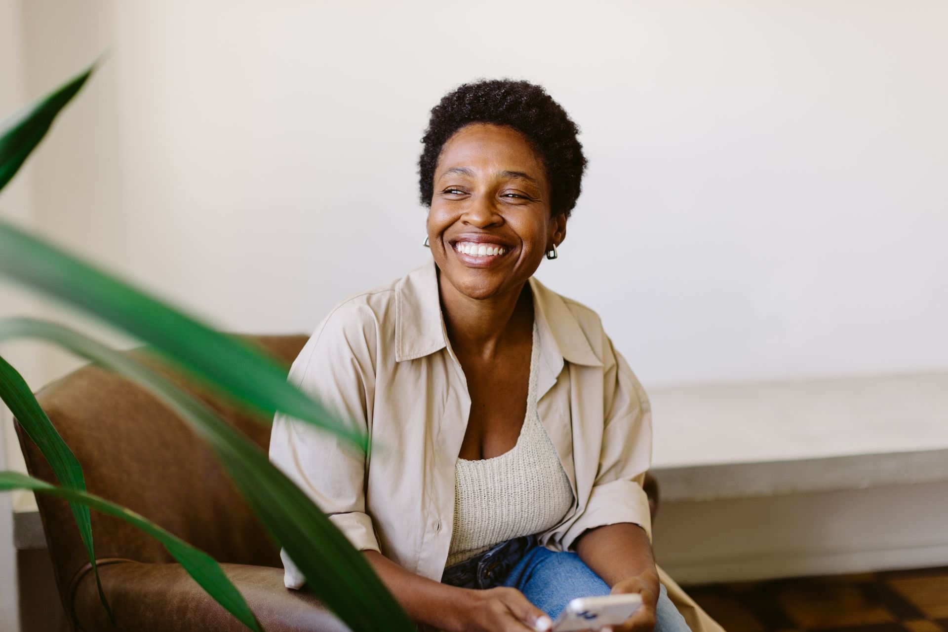 Woman smiling, sitting on a brown couch, holding a phone, wearing a light-colored shirt and jeans.