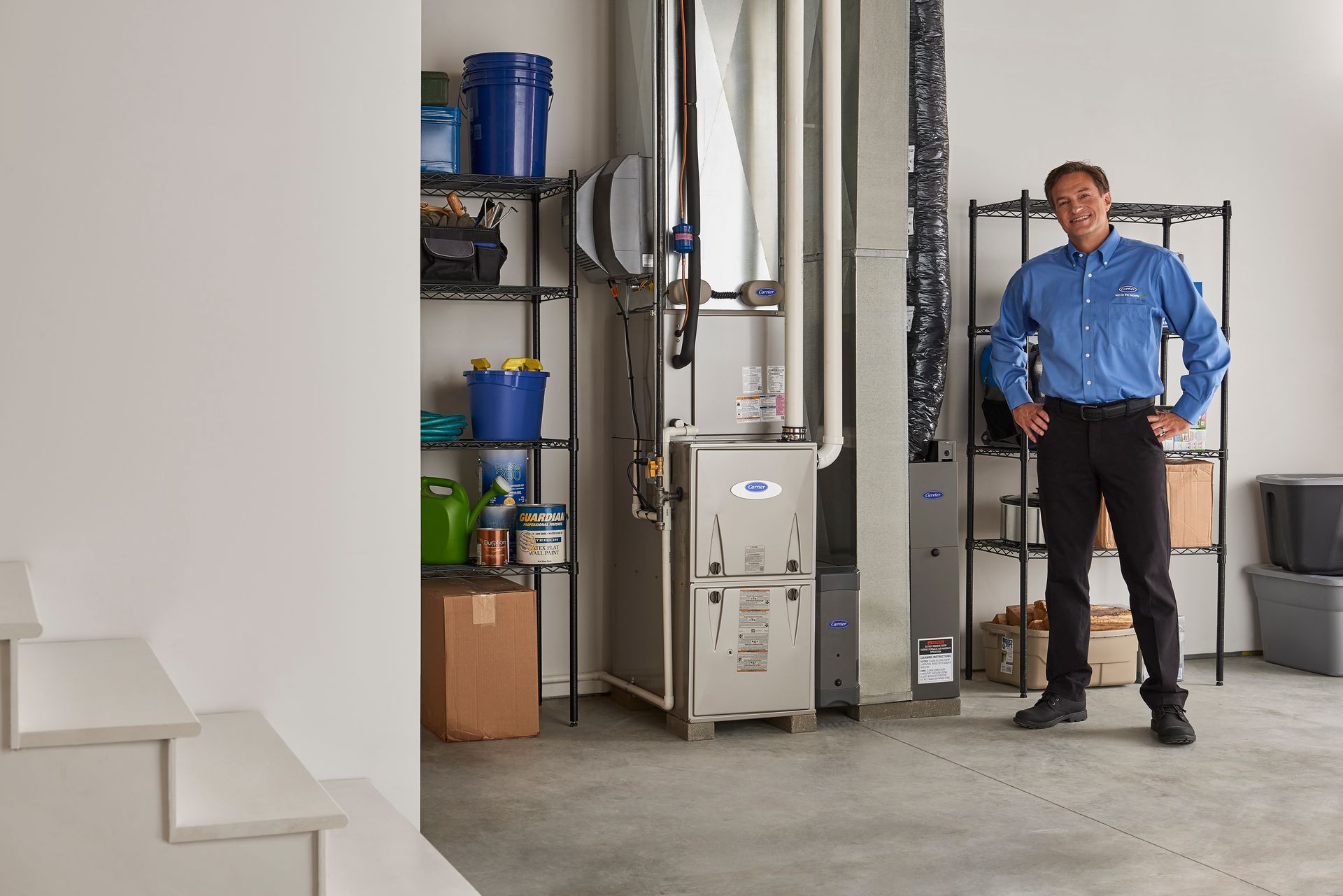 HVAC technician standing in a utility room next to furnace. White walls, storage shelves, and stairs.