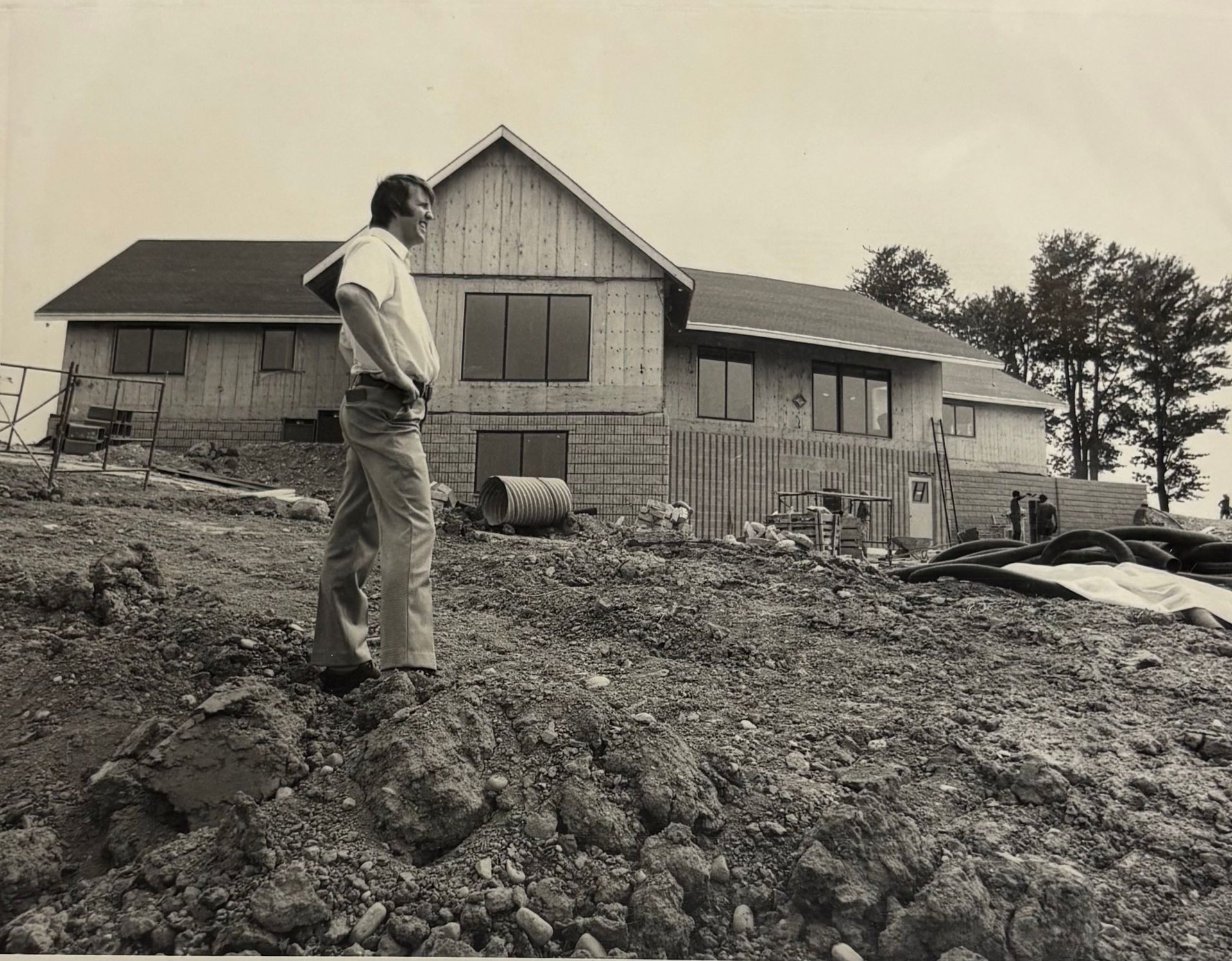 A person stands on a dirt slope in front of a large, unclad house under construction with exposed wooden framing.
