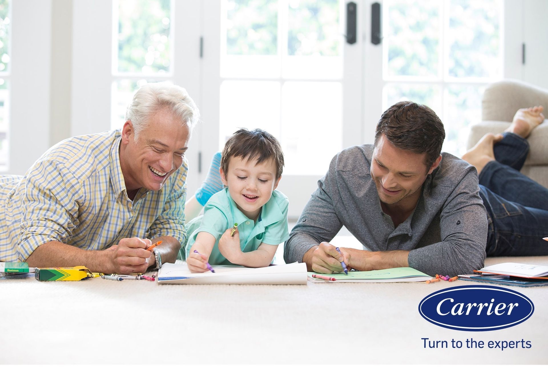 Three people smiling and coloring together on a living room floor, with a Carrier logo in the bottom right corner.