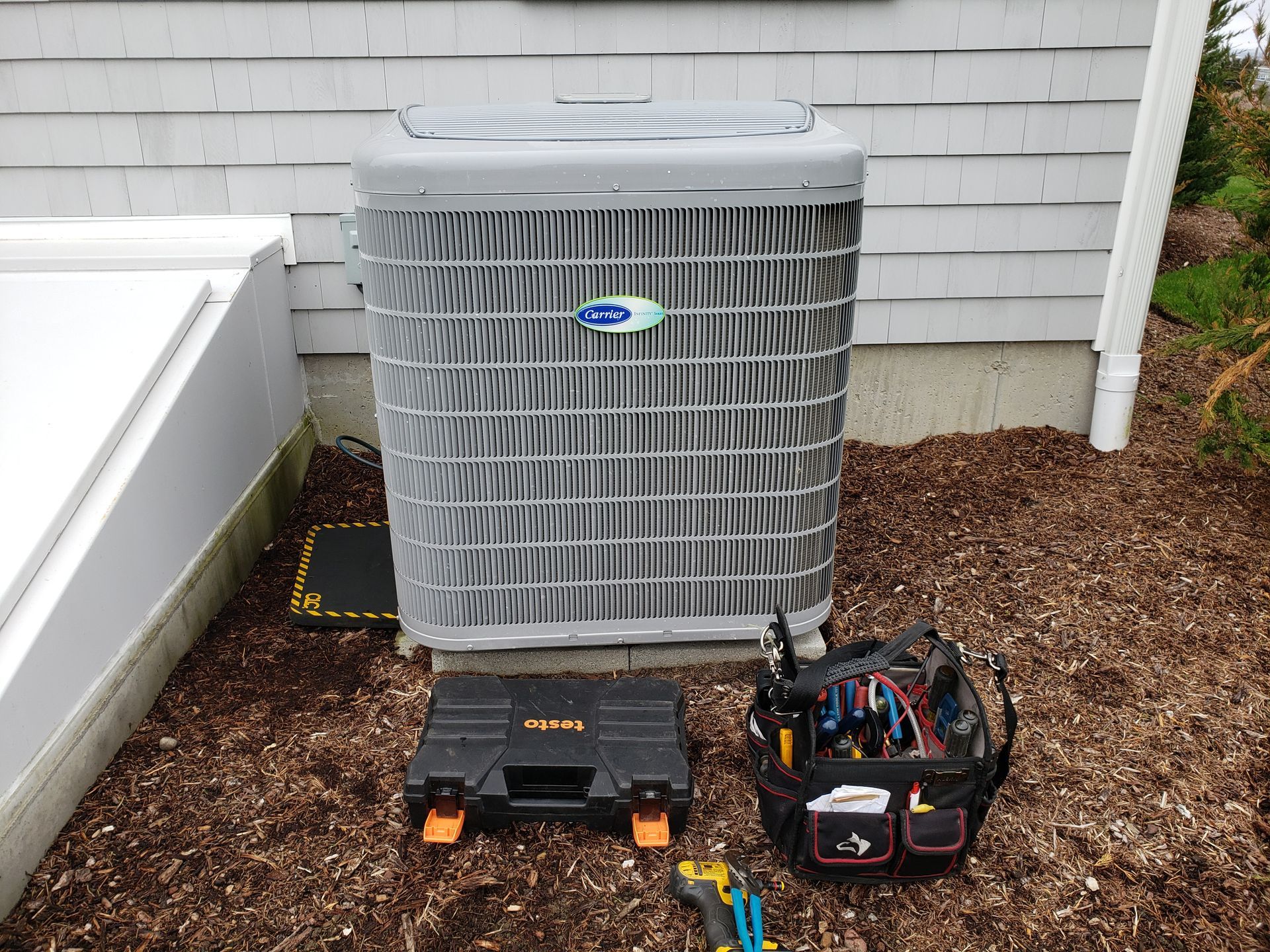 Air conditioner unit with tools nearby, set against a light-colored building, outdoors on mulch.