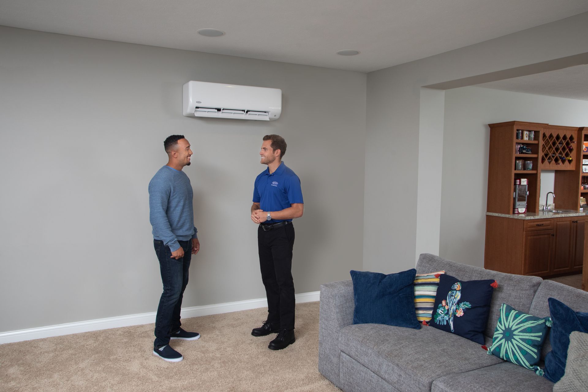 Two people stand in a living room, talking under a wall-mounted ductless air conditioning unit.