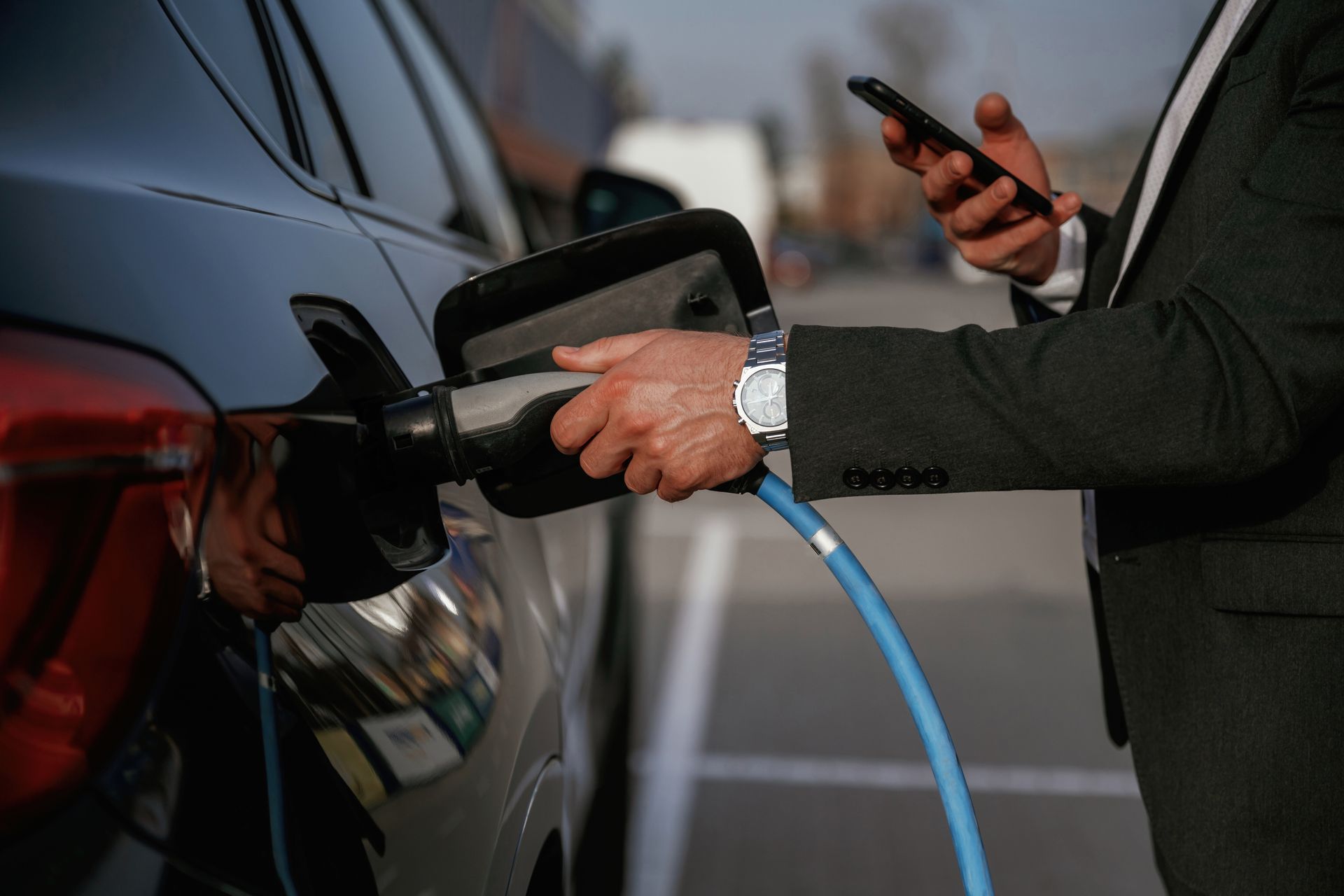 A person in a business suit plugs a charging cable into an electric vehicle while looking at a smartphone.