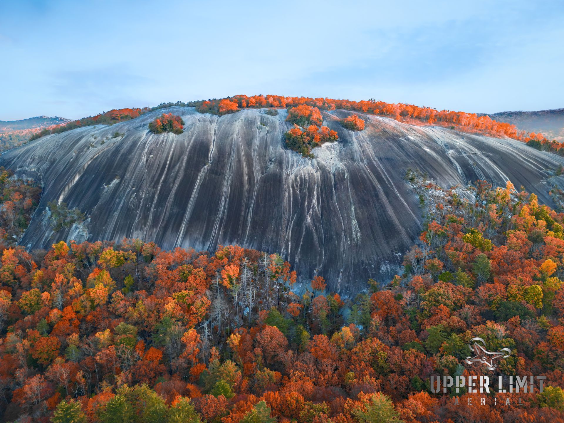 Large, gray granite dome with fall foliage in shades of orange and red at the base and top, under a blue sky.