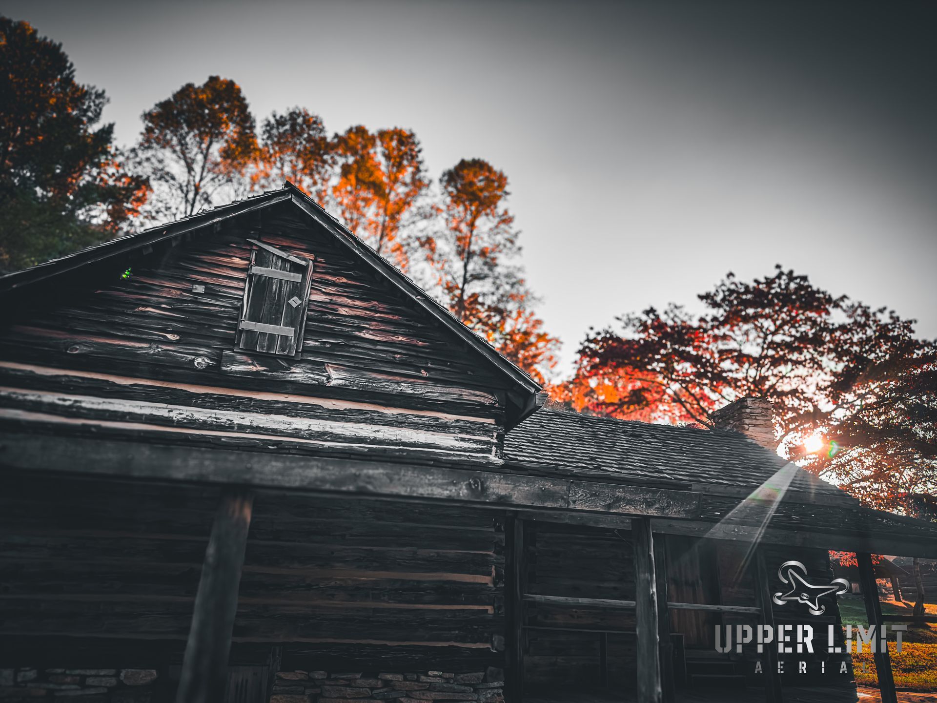 Old wooden cabin in a forest with autumn-colored trees and the setting sun.