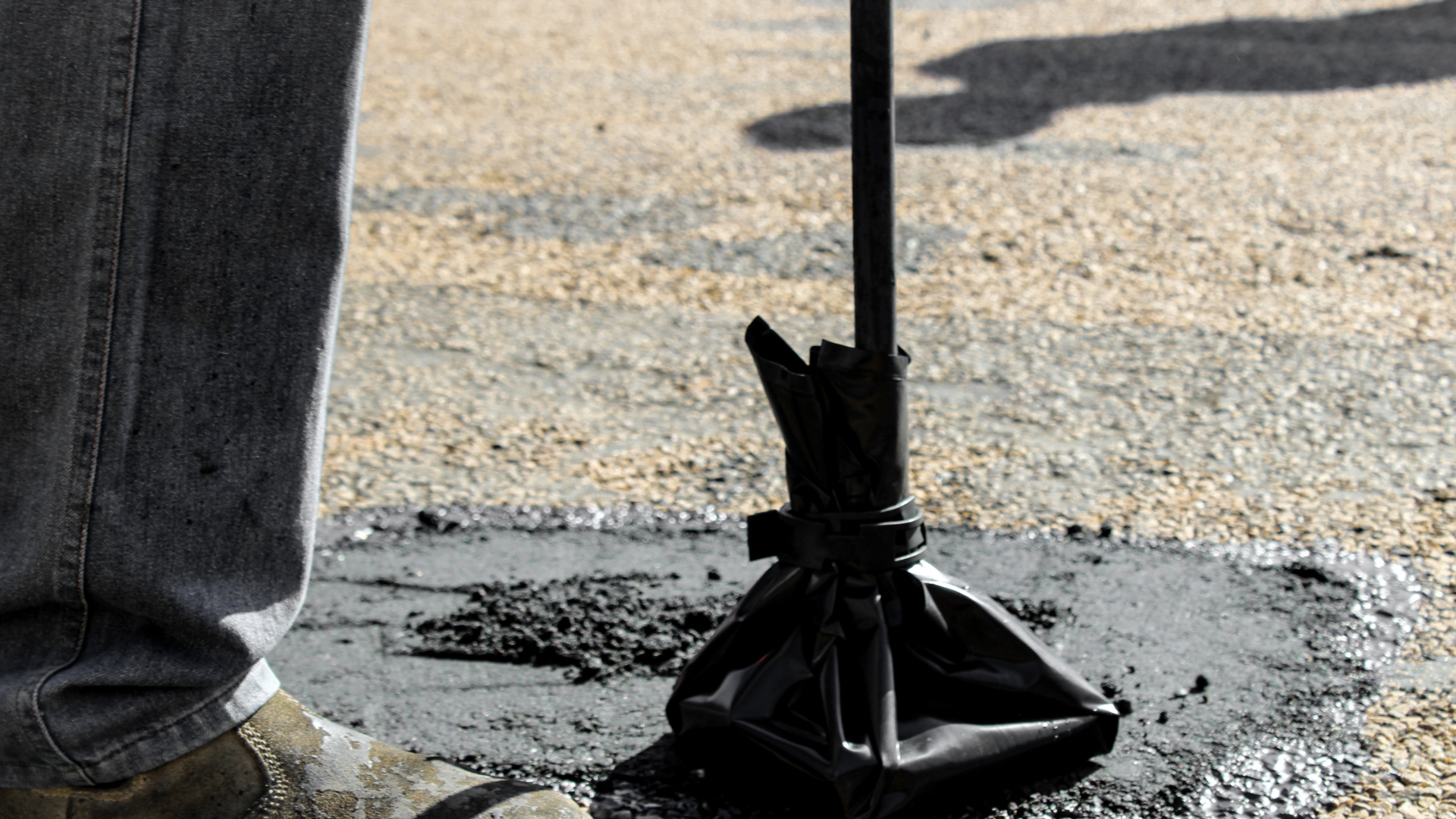 Person's legs and shoe near a black, filled asphalt crack repair bag and tool on a cracked asphalt surface.