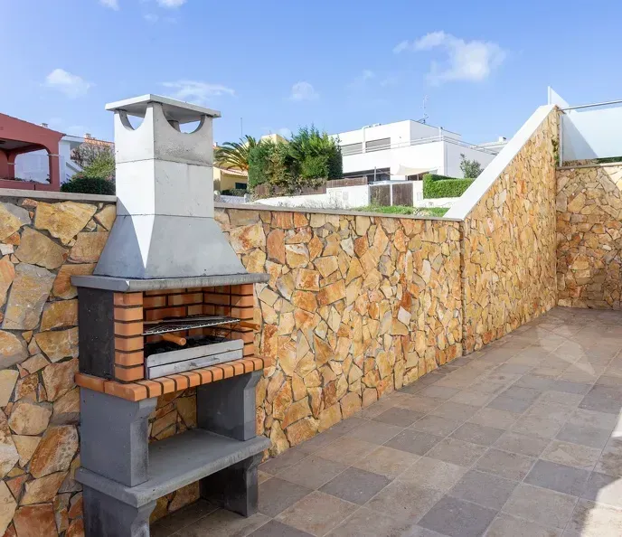 Brick barbecue grill set against a stone wall and tiled patio, under a sunny blue sky.