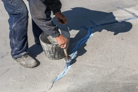 Person filling a crack in concrete with blue sealant. Bucket and brush are present.