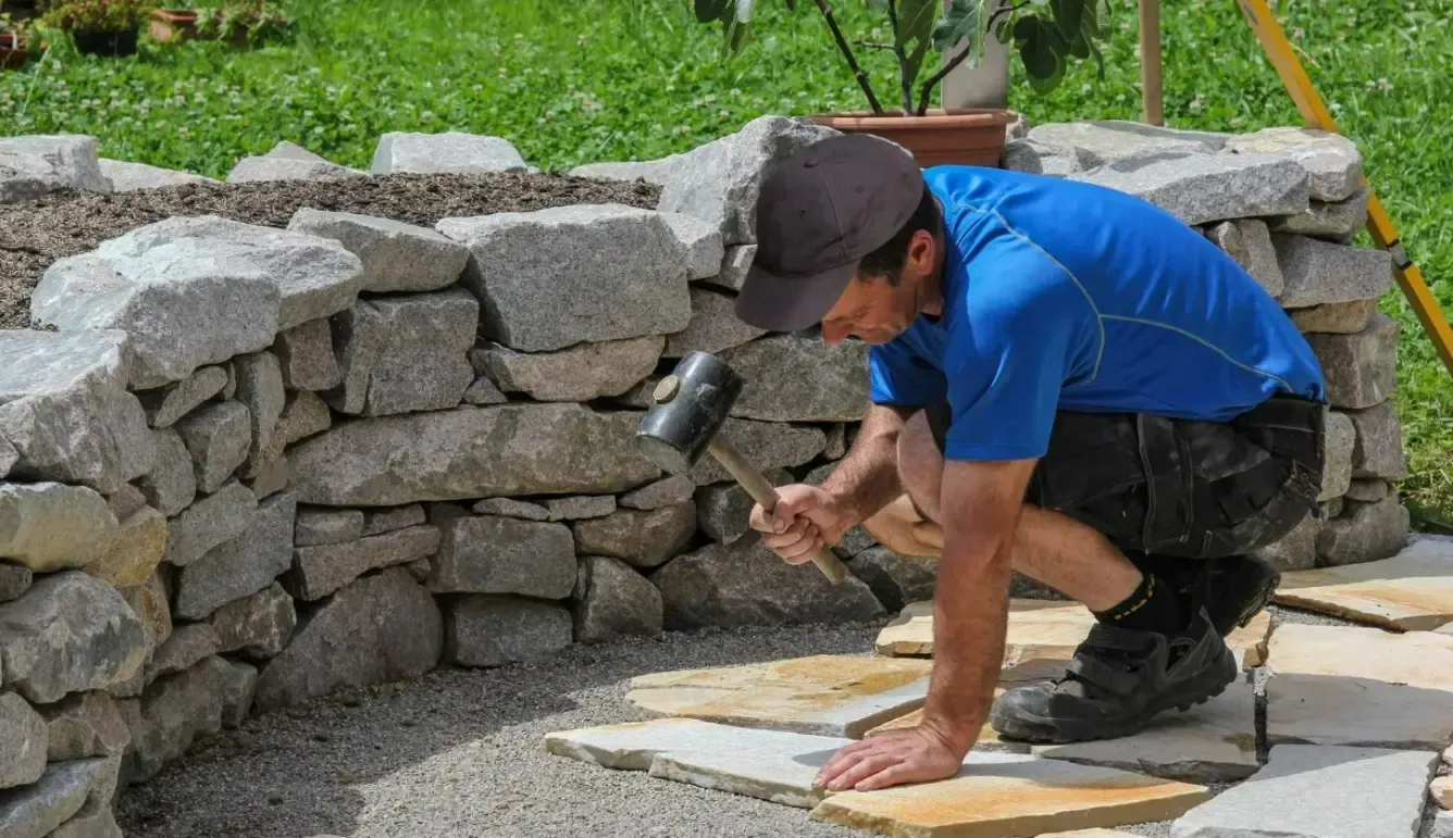 Man building a stone wall and laying pavers outdoors, using a mallet.