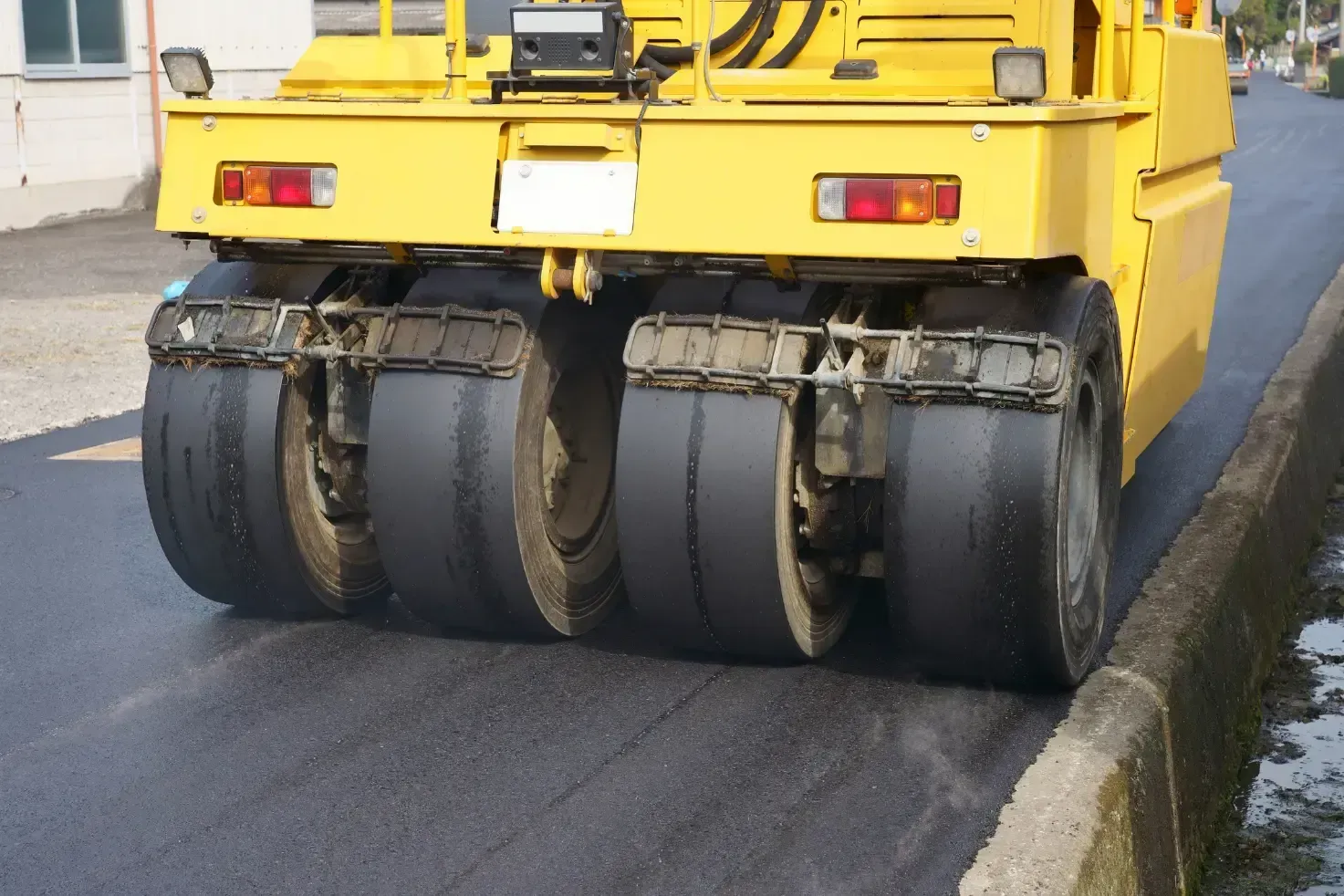 Yellow asphalt roller compacting fresh asphalt next to a curb.