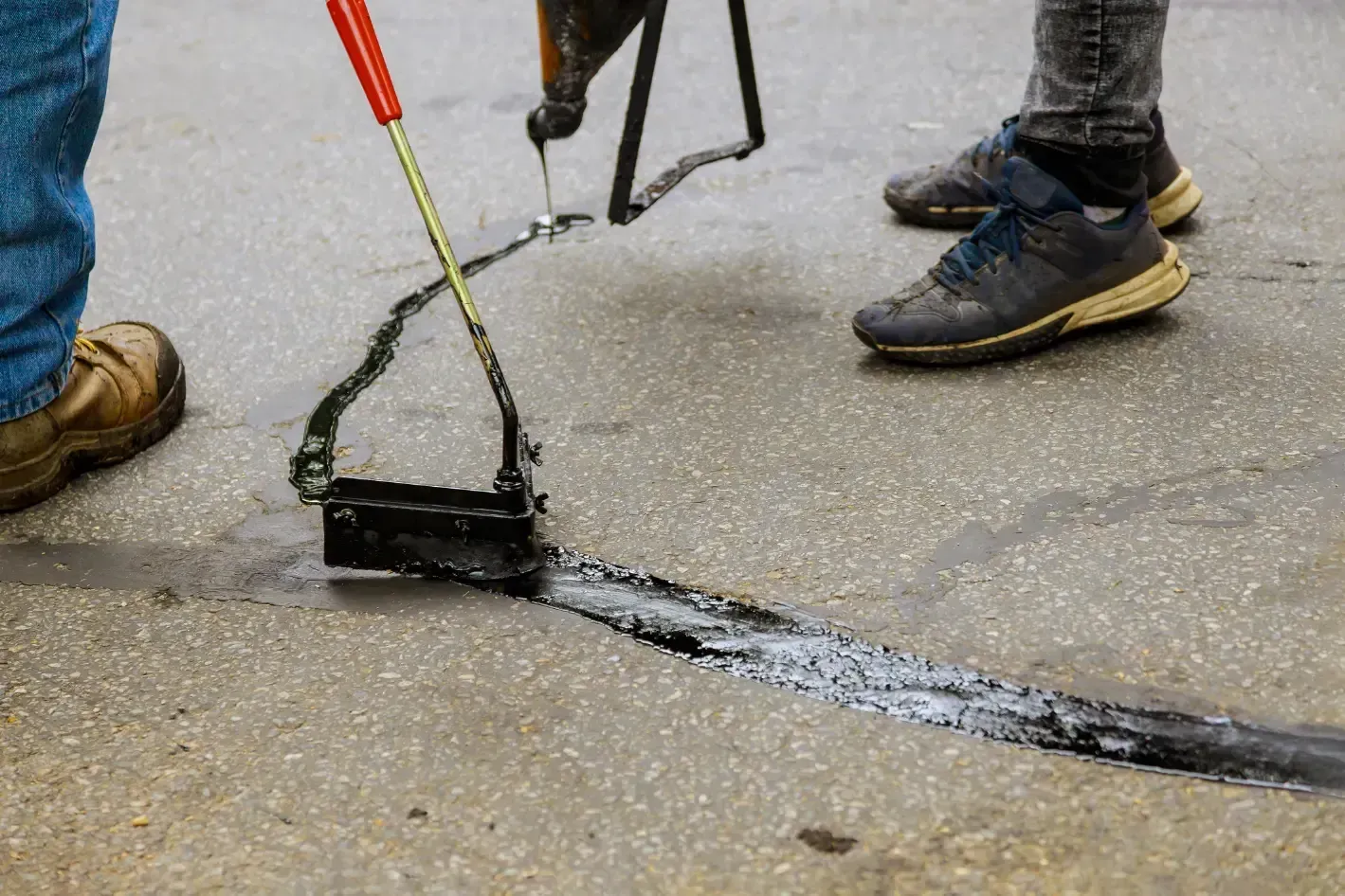 Two people repairing a crack in pavement with a black sealant.