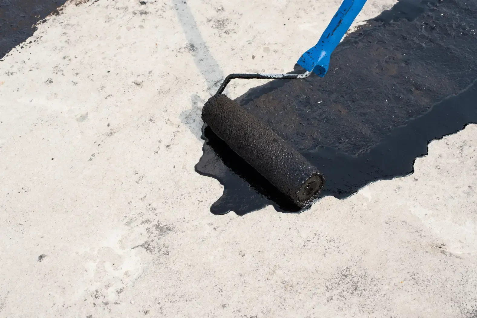 A person using a paint roller to apply black sealant to a light-colored surface.
