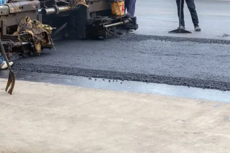 Road paving in progress: asphalt laid by a machine, smoothed by a worker; concrete sidewalk in foreground.