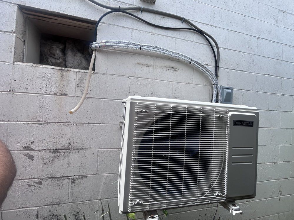 Air conditioner unit mounted on a brick wall, pipes and cables entering a window opening.