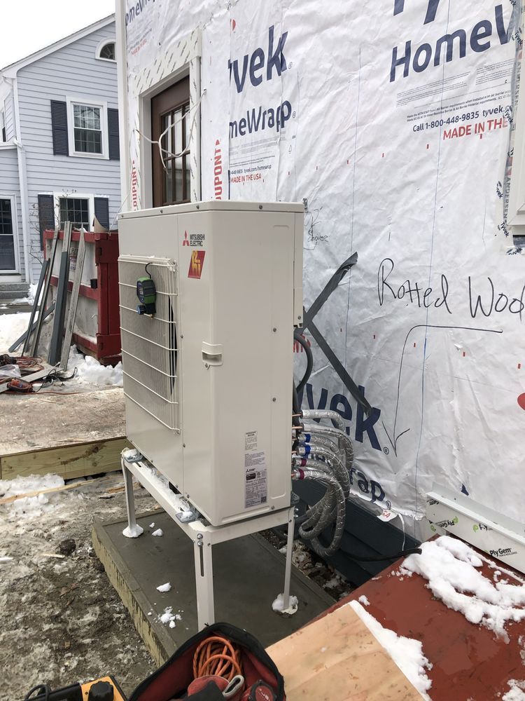 White heat pump unit mounted on a stand next to a house with exposed siding in a snowy outdoor setting.