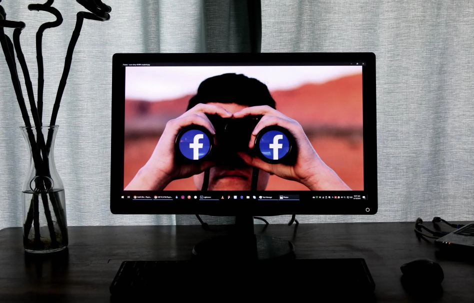 Computer monitor displaying a person looking through binoculars with Facebook logos; setting indoors.