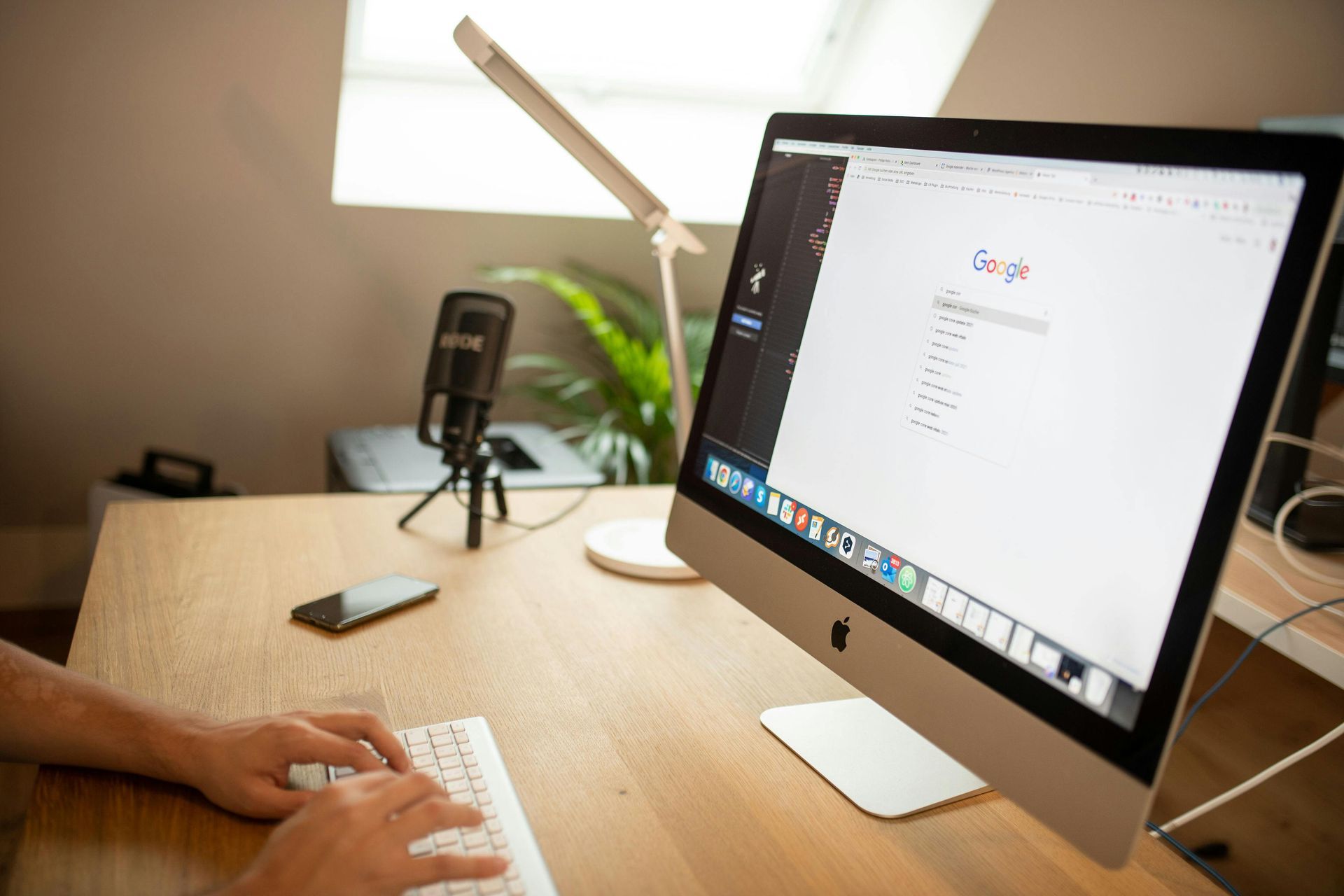 Person typing on keyboard, Google search on screen, desk setup with microphone and lamp.