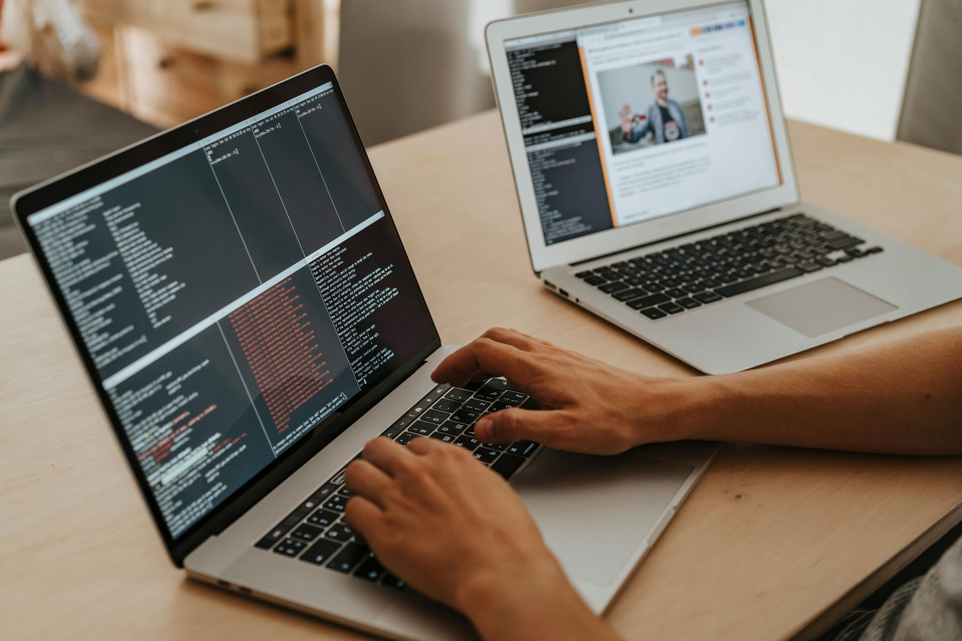 Hands typing on a laptop with code displayed, another laptop shows a webpage, on a light wood table.