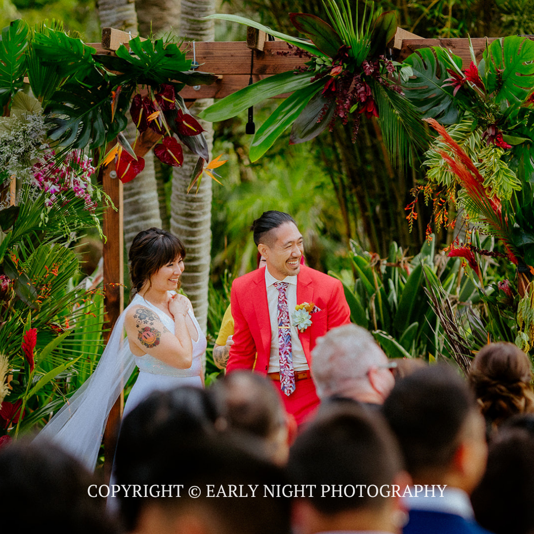 Outdoor wedding ceremony with bride in white and groom in red amid tropical greenery