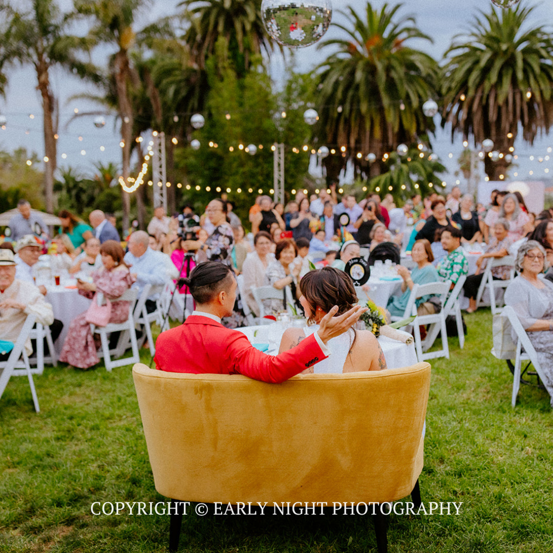 Couple seated in a gold chair at an outdoor wedding reception with guests, palm trees, and string lights.