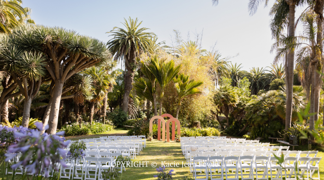 Outdoor wedding setup with white chairs facing a floral arch in a lush garden