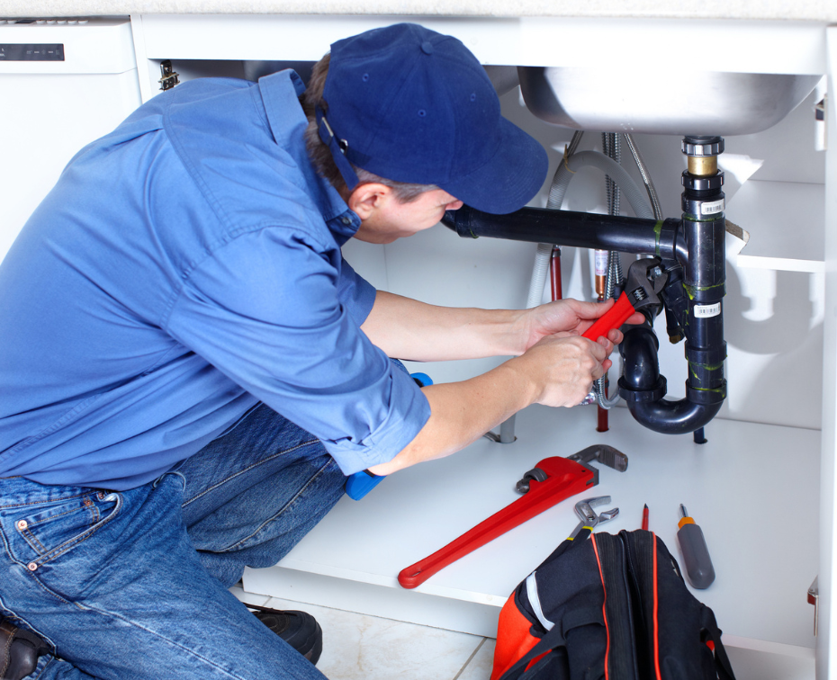Plumber in blue shirt and cap, crouched under a kitchen sink, using a wrench to repair pipes.