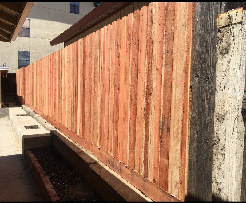 Wooden fence of vertical planks, reddish-brown, along a concrete structure with a building in the background.