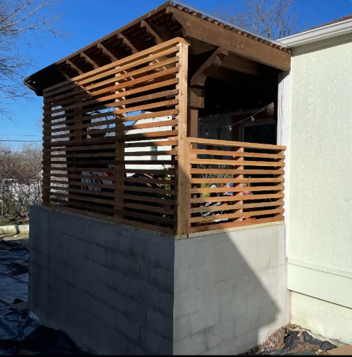 A wooden pergola with slatted walls, built atop a concrete foundation next to a light-colored building.